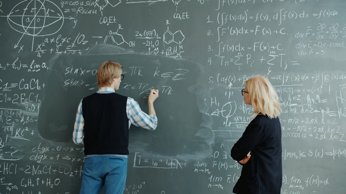 Student writes math equations on blackboard while teacher watches.